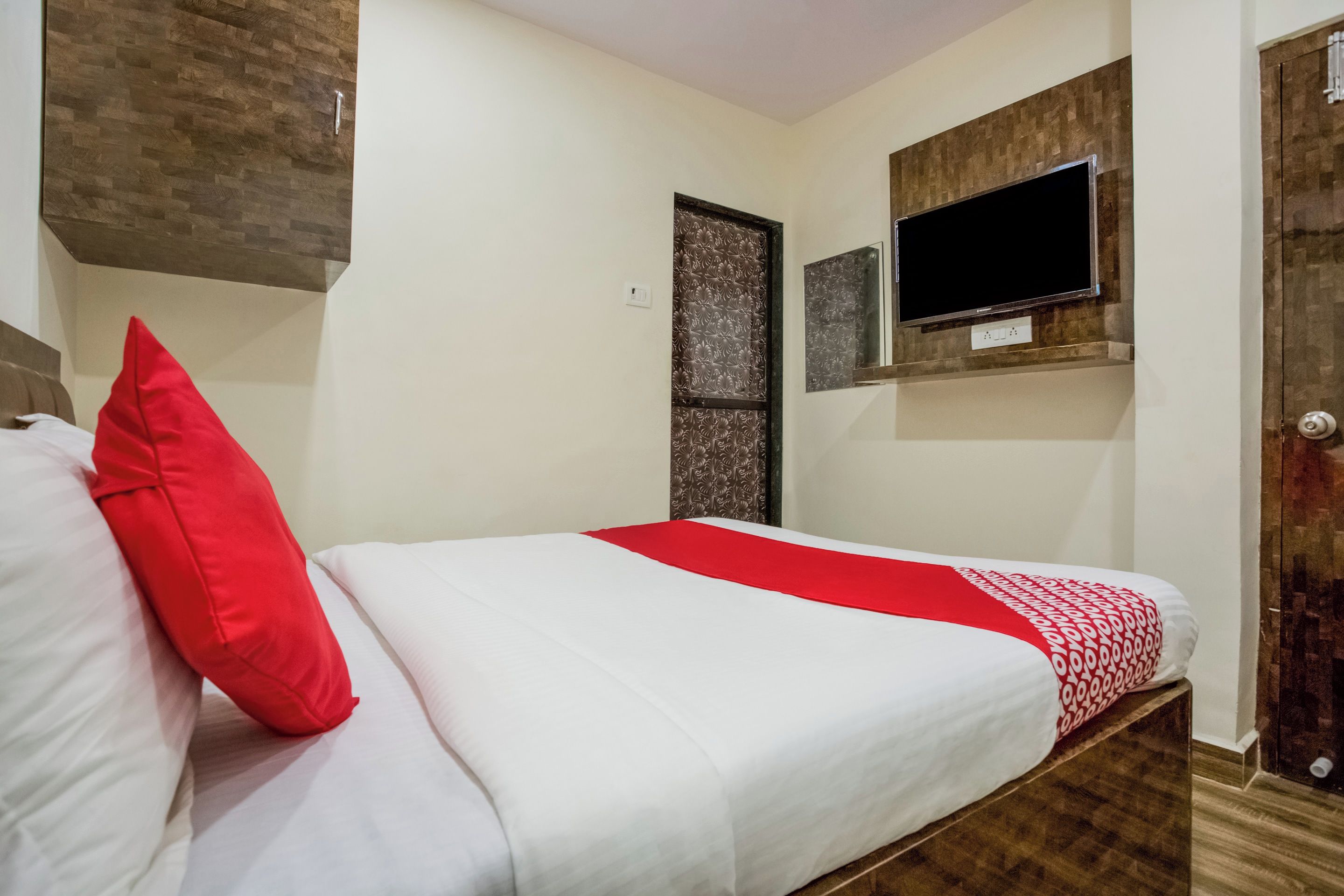 A bedroom with a bed, a red pillow, a television mounted on the wall, and a door with a patterned design.