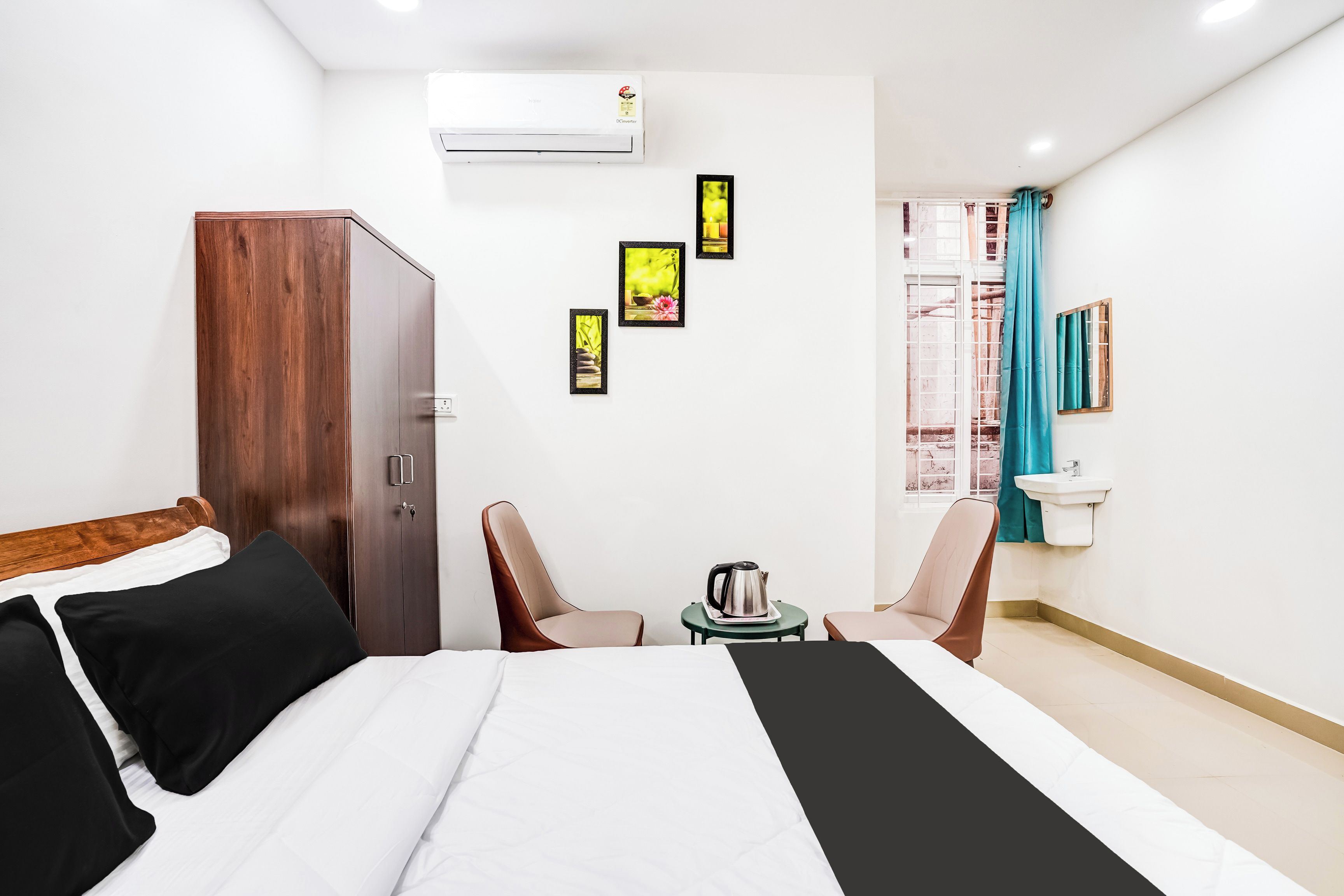 Habitación de hotel con cama, armario, escritorio con tetera, silla, lavamanos y ventana con cortinas.