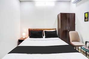 A hotel room with a bed, a wardrobe, a chair, a nightstand with a lamp, and a small table with a kettle. - Hotel O HMR Inn (Bangalore)