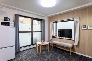 Et kompakt hotelværelse med tekøkken, et lille rundt bord, en bænk og et stort vindue med gardiner.
