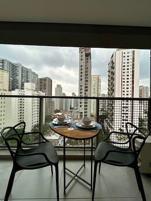 A balcony with two chairs, a table set with plates, bowls, and glasses, overlooking a cityscape with high-rise buildings. - Level Brooklin 1217 (São Paulo)