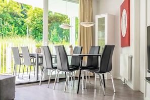 A dining area with a table and chairs, a television, and a view of greenery outside. - Dune Villa 02, Apartment 02 (Lübeck-Travemünde)