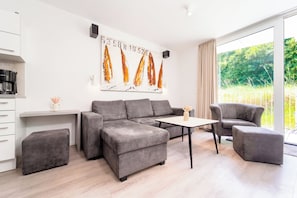 A modern living room with a grey sofa set, a coffee table, and a dining table. There is a kitchen area with a coffee machine and cabinets. A large window provides a view of greenery outside. - Dune Villa 02, Apartment 02 (Lübeck-Travemünde)