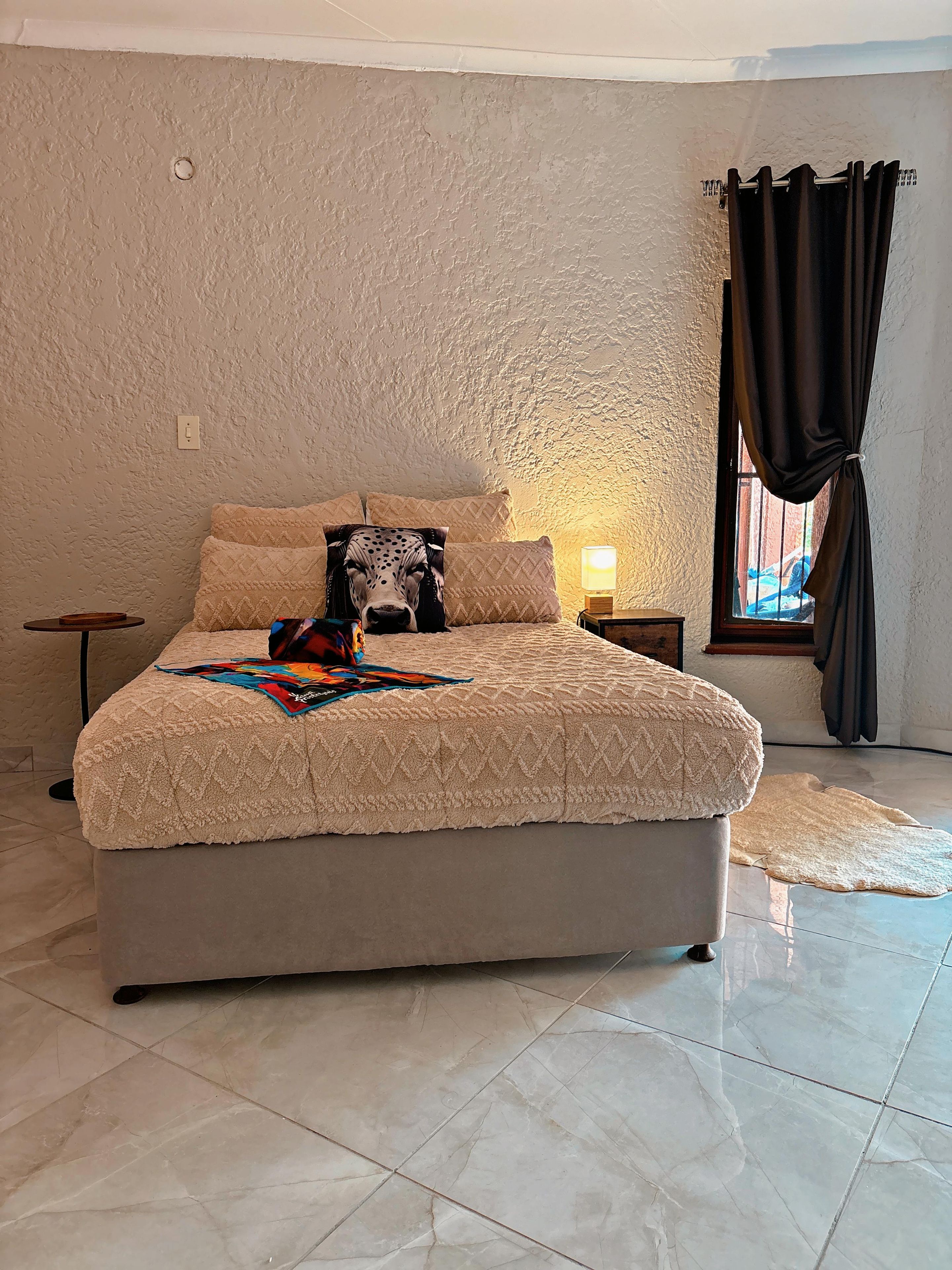 Un dormitorio con cama, mesita de noche, lámpara, ventana con cortinas y piso de baldosas.