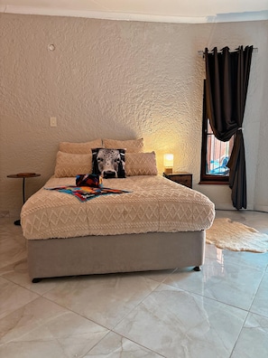 Un dormitorio con cama, mesita de noche, lámpara, ventana con cortinas y piso de baldosas.