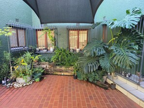 Un patio con piso de ladrillo, plantas en macetas, un pequeño elemento decorativo de agua y una cerca de metal.