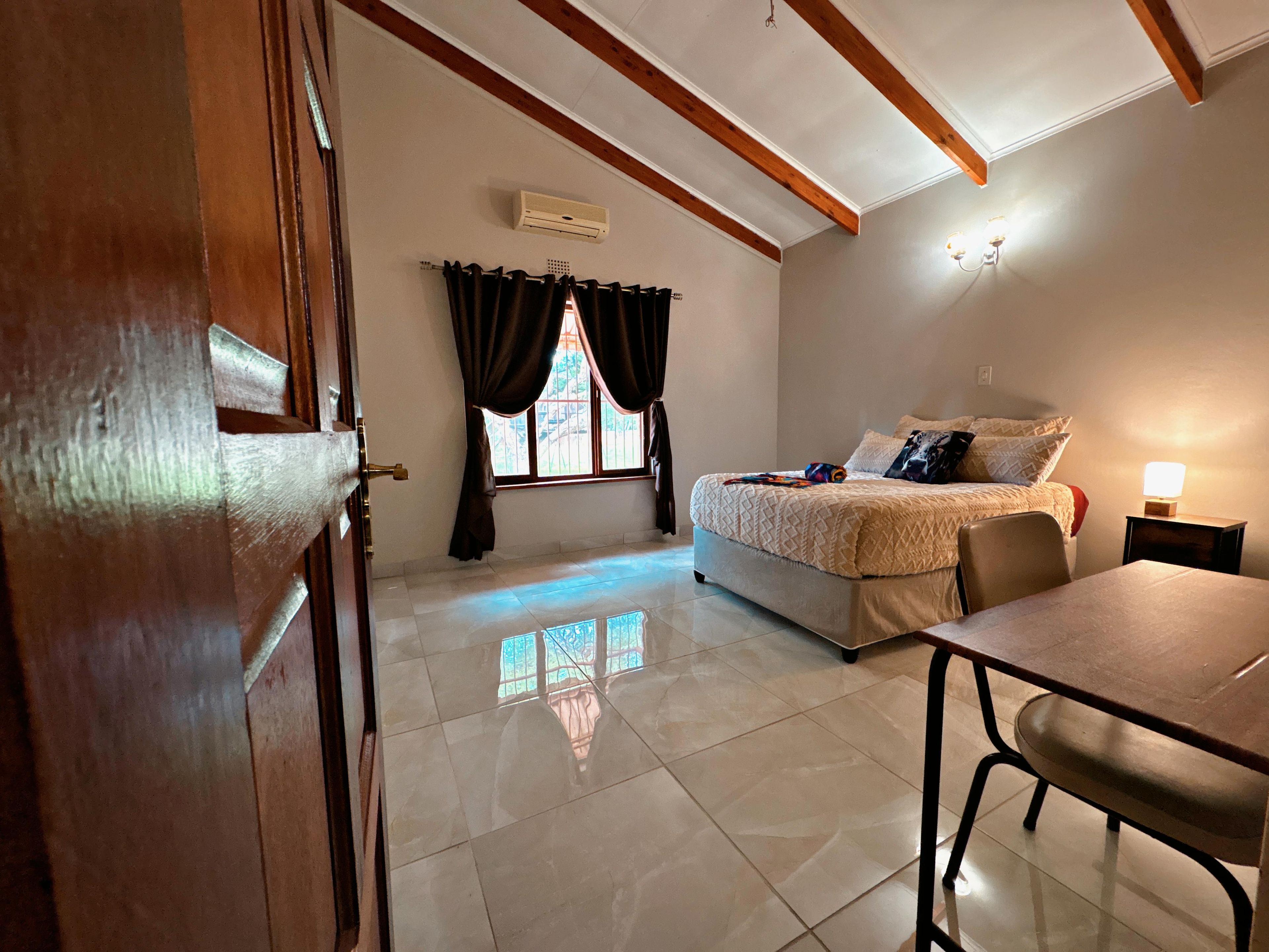 A bedroom with a bed, a desk, and a chair. There is a window with curtains, an air conditioning unit, and a lamp on a bedside table.