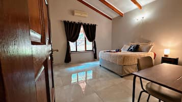 A bedroom with a bed, a desk, and a chair. There is a window with curtains, an air conditioning unit, and a lamp on a bedside table.