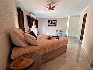 A bedroom with a bed, pillows, a wooden side table, a ceiling fan, a window with curtains, and a wall-mounted picture.
