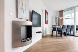 A modern living room with a flat-screen TV mounted on the wall, a built-in fireplace, a dining area with a table and chairs, and a large window with curtains. - Dune Villa 02, Apartment 03 (Lübeck-Travemünde)