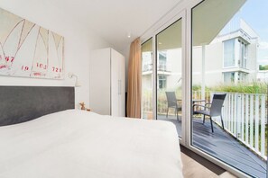 A modern bedroom with a large bed, a wardrobe, and a balcony with a view of neighboring buildings. - Dünenvilla 18, Apartment 02 (Lübeck-Travemünde)