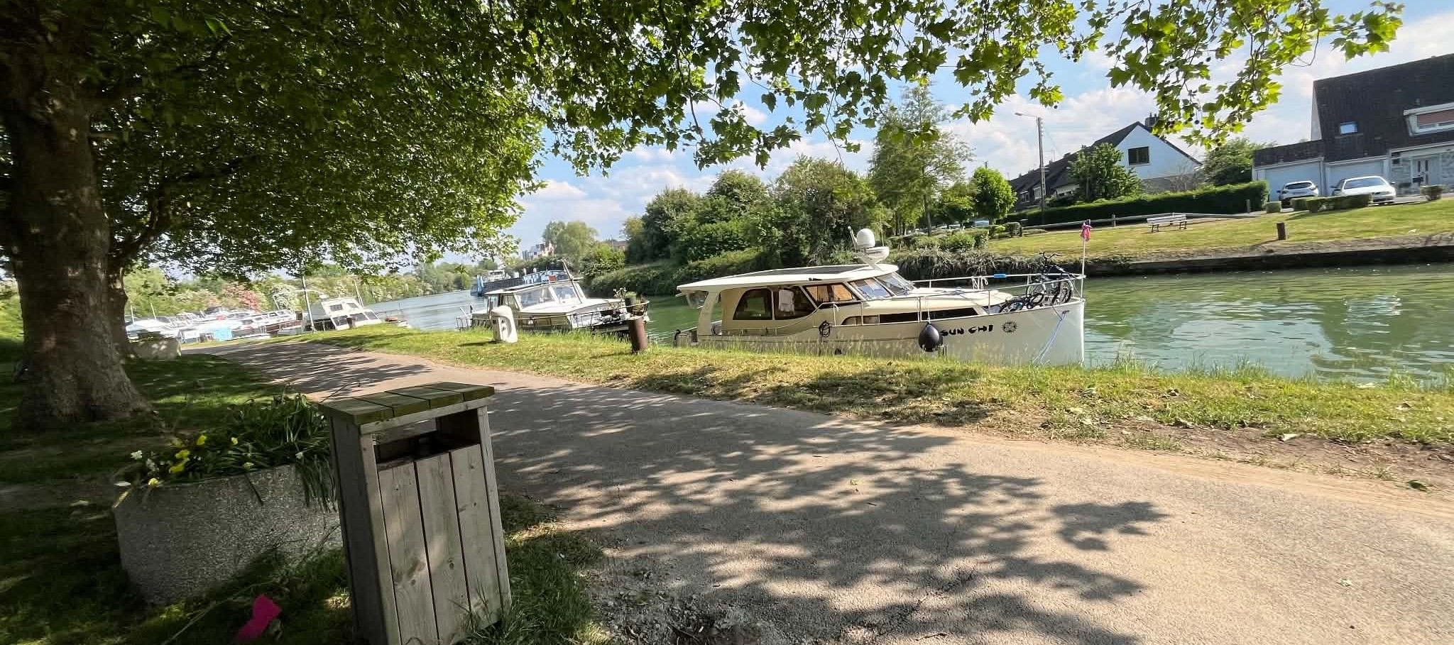 A riverside path with boats docked, a tree-lined area, and a building in the background.