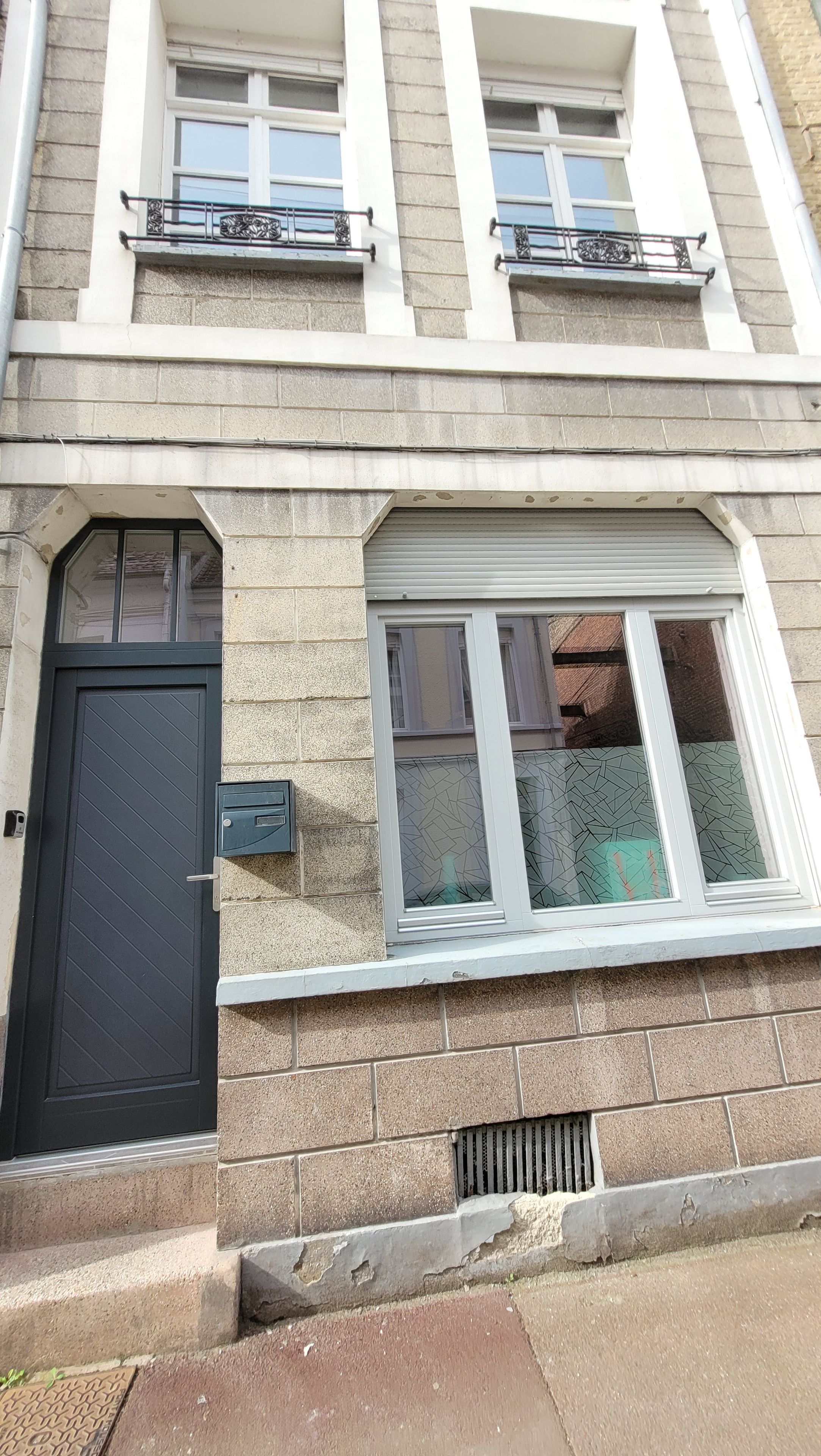 Un bâtiment avec une porte noire, une boîte aux lettres et une fenêtre ornée d’une grille métallique.