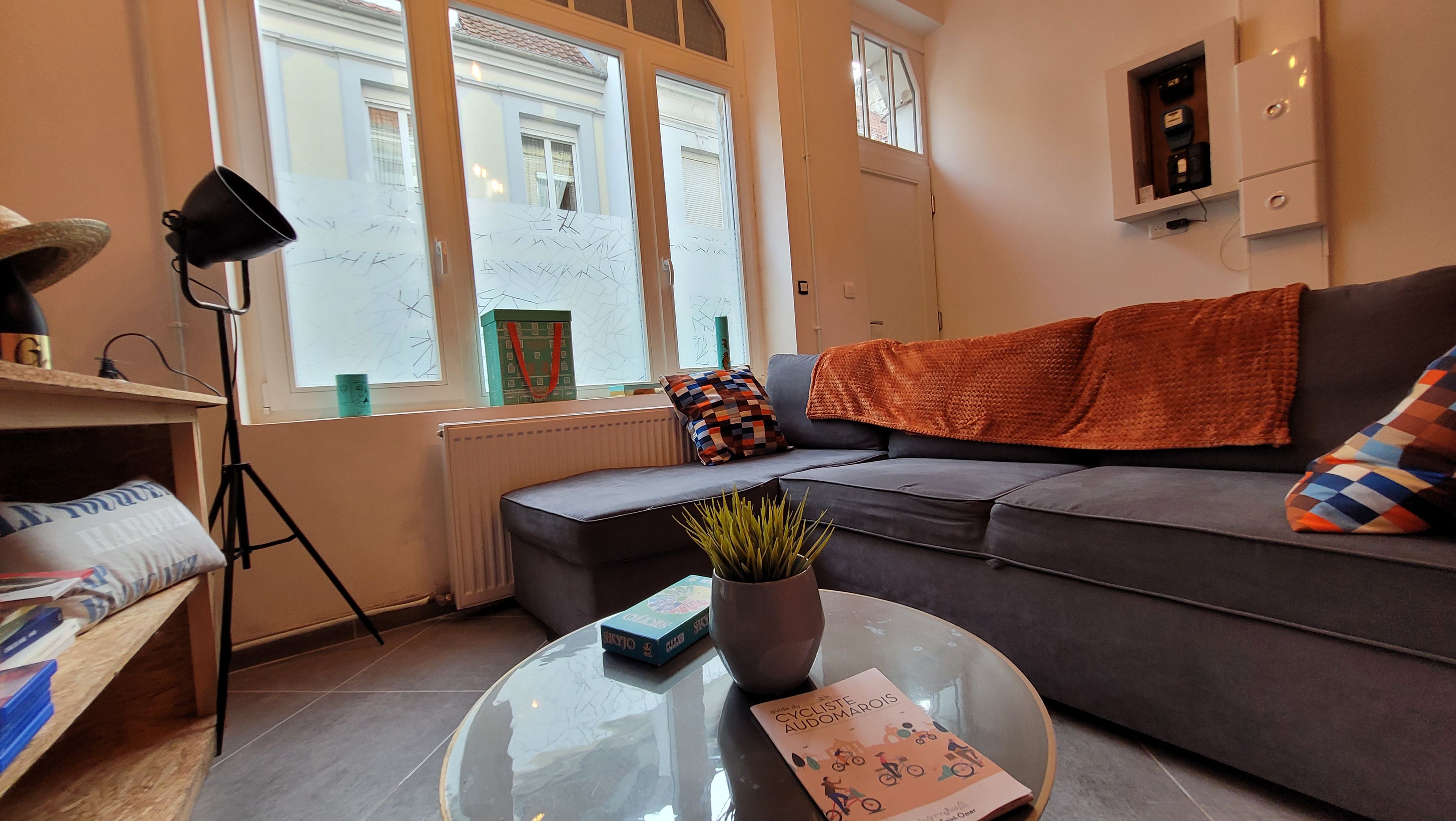 A modern living room with a sofa, a coffee table with a book, a floor lamp, and a view of the street through large windows.
