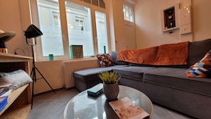 Ein modernes Wohnzimmer mit einer Couch, einem Couchtisch mit einem Buch, einer Stehlampe und Blick auf die Straße durch große Fenster.