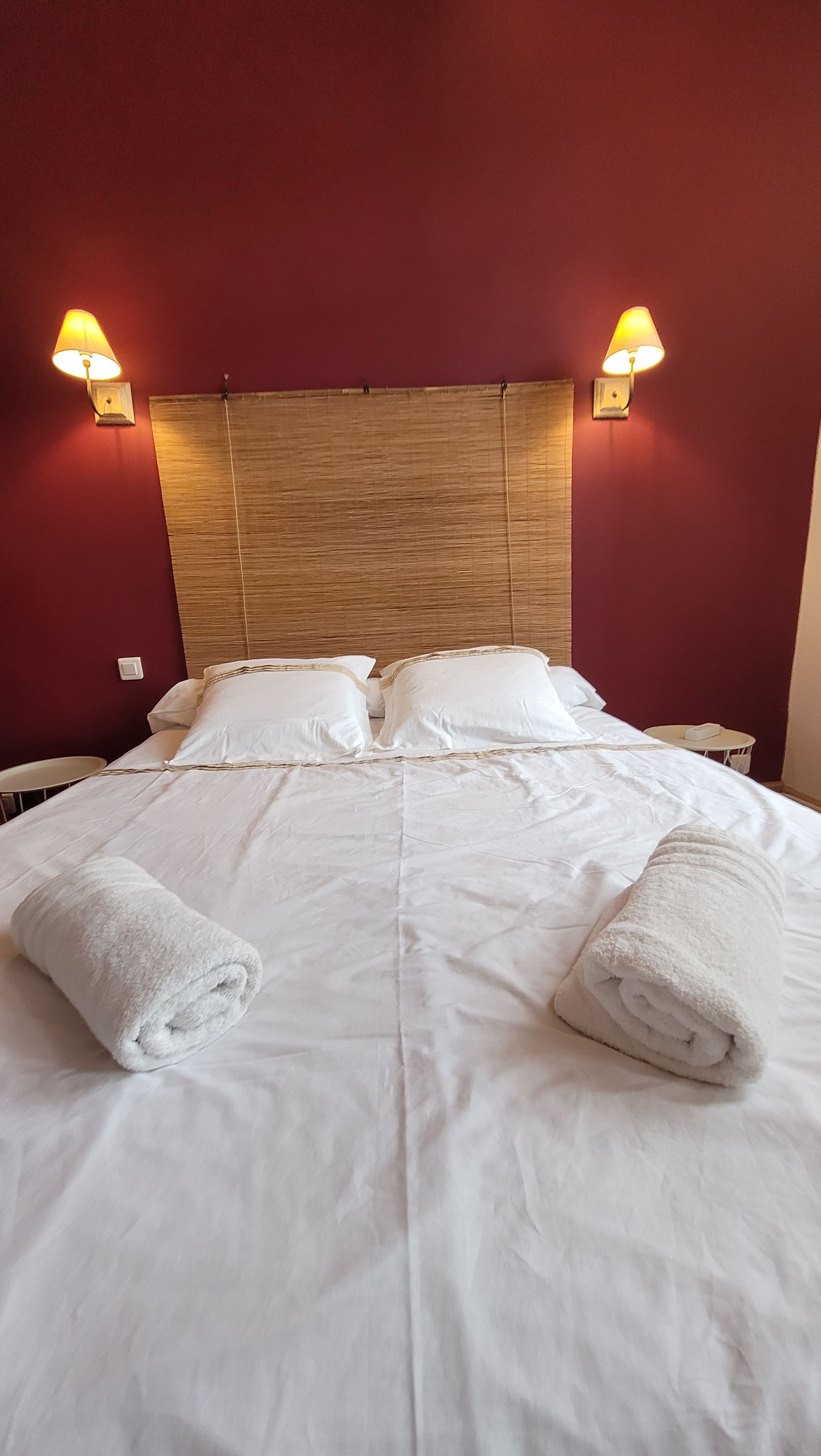 Un lit bien fait, avec du linge de lit blanc et deux serviettes enroulées, une tête de lit en bois et deux lampes fixées au mur.