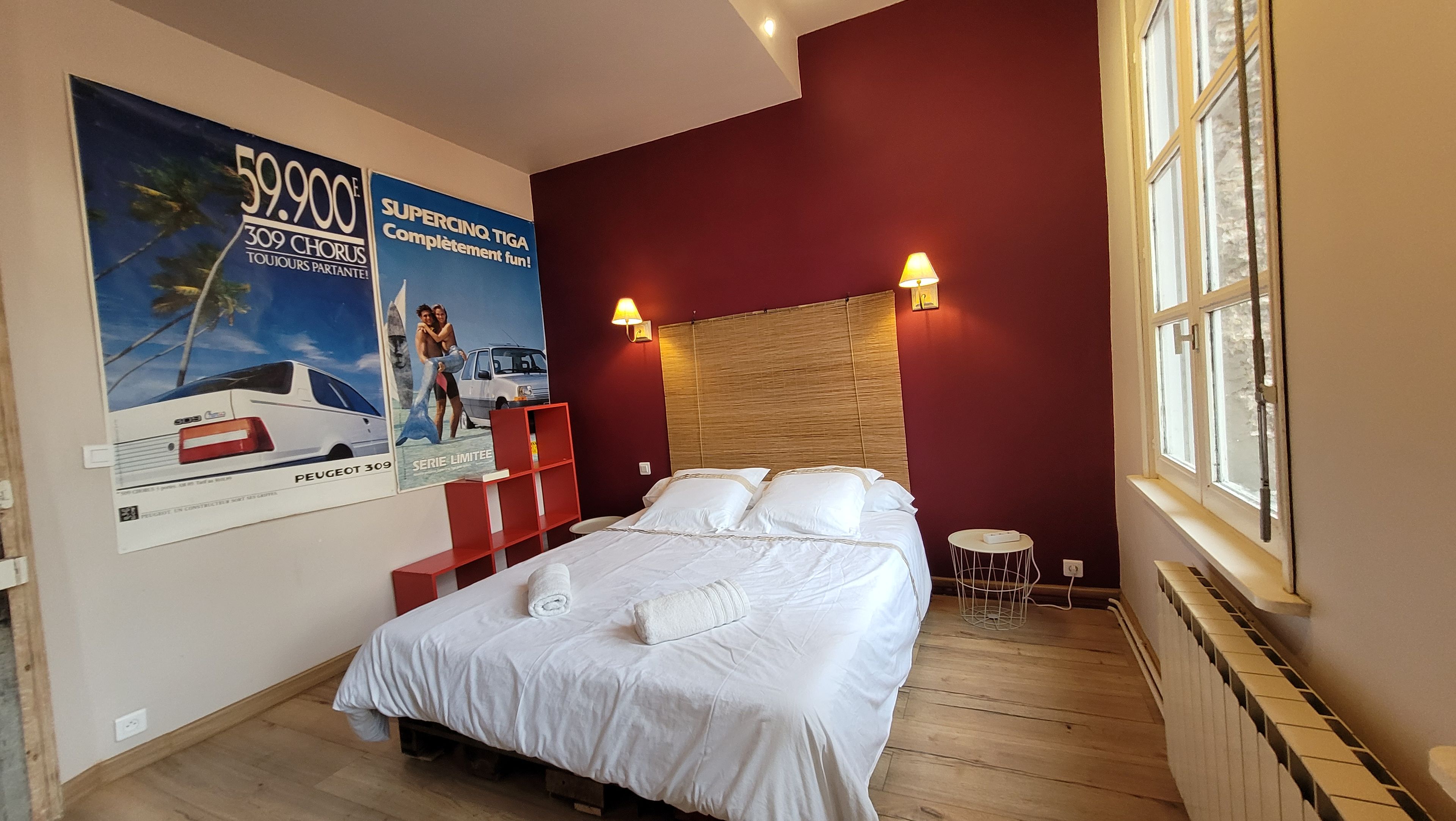 Ein Hotelzimmer mit einem Bett, einem Nachttisch, einer roten Ablage, einem Poster an der Wand und einem Fenster.