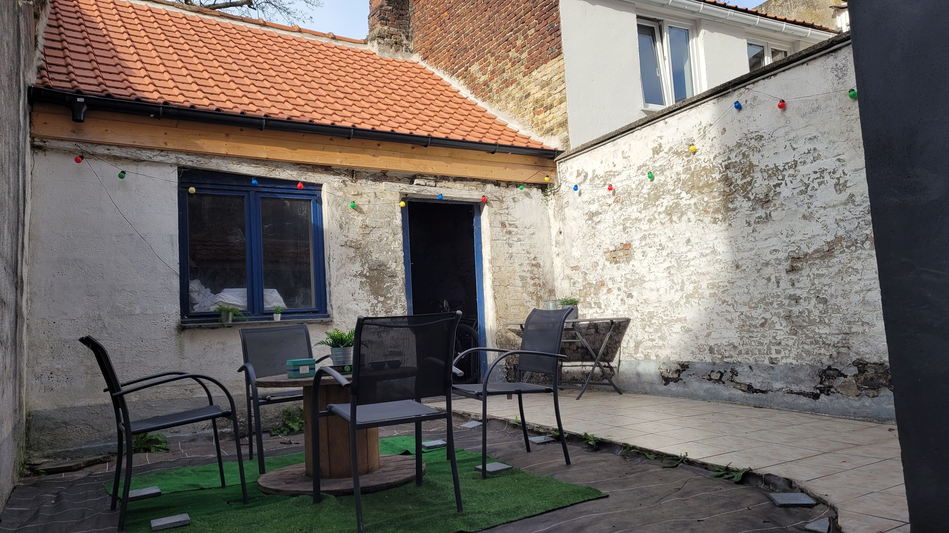 Une cour avec un toit en tuiles, une table en bois entourée de chaises et un petit jardin.