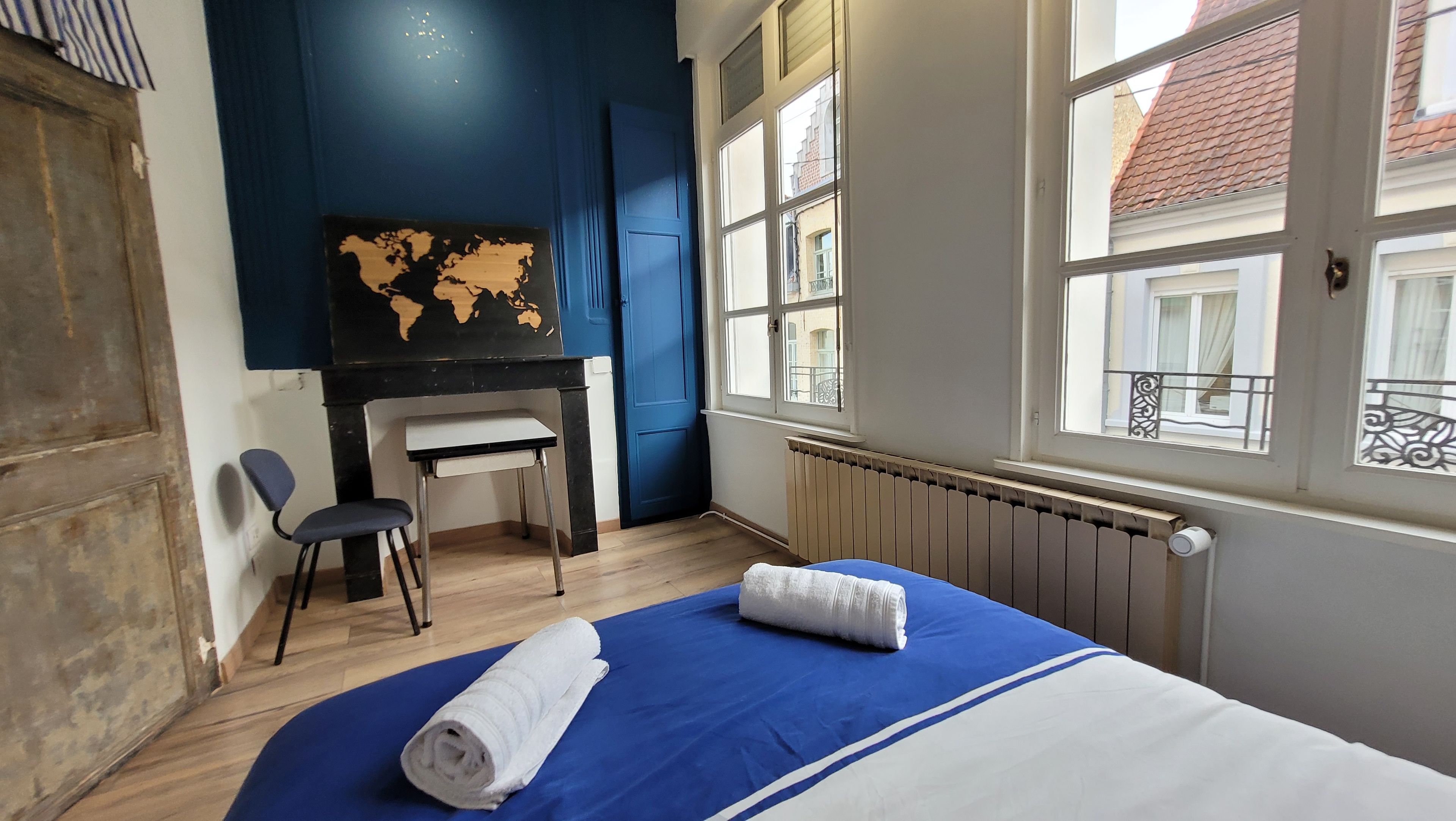 Ein Zimmer mit einem Bett, einem Schreibtisch, einem Stuhl, einer Karte an der Wand und einem Fenster mit Blick auf Gebäude.