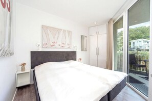 A modern bedroom with a large bed, a wardrobe, a bedside table, a wall-mounted artwork, and a view of the outdoors. - Dünenvilla 18, Apartment 01 (Lübeck-Travemünde)