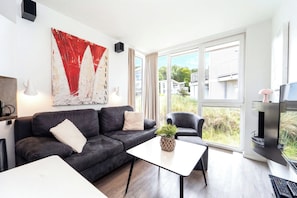 A modern living room with a dark sofa, a white coffee table, and a large abstract painting. - Dünenvilla 18, Apartment 01 (Lübeck-Travemünde)