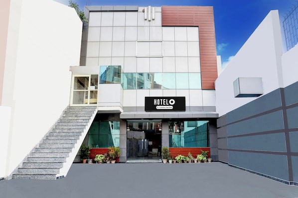 Un hôtel moderne, reconnaissable à son grand panneau lumineux indiquant « HÔTEL », avec un escalier menant à l’entrée et des plantes en pot à sa base.