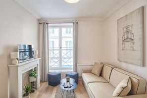 A living room with a sofa, ottomans, a fireplace, a television, and a large window. - Lovely apartment with AC - Center of Paris (Paris)