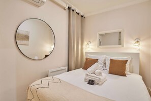 A hotel room with a bed, a round mirror, a headboard with two pillows, a wall-mounted light, and a window with curtains. - Lovely apartment with AC - Center of Paris (Paris)