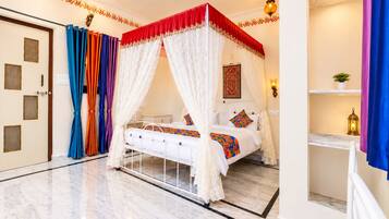 Una cama con dosel y cuatro columnas, con cubrecama blanco y almohadas de colores, suelo de mármol y un ventilador de techo decorativo.