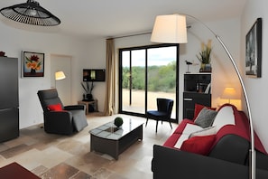 Ein modernes Wohnzimmer mit Sofa, Sessel und Couchtisch. Ein großes Fenster bietet Blick auf Bäume, und eine Tür führt nach draußen.