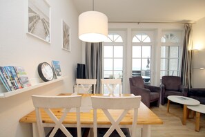A room with a wooden table and chairs, a bookshelf with magazines, a clock, and a large window with curtains. - 2-room vacation rental, approx. 37 m², located right on the beach with an ocean view (Wyk auf Föhr)