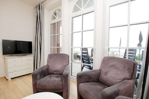 A living room with two armchairs, a TV on a cabinet, and large windows with a view of the outside. - 2-room vacation rental, approx. 37 m², located right on the beach with an ocean view (Wyk auf Föhr)
