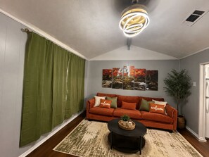 A living room with a sofa, a coffee table, a green curtain, a wall with a waterfall painting, and a ceiling light fixture. - Orange Haven By Kay Mac Rentals (Meridian)
