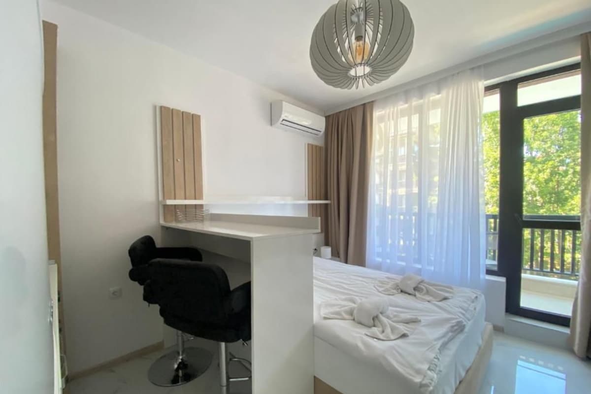 A hotel room with a bed, a desk with a chair, an air conditioning unit, and a view of greenery through the window.