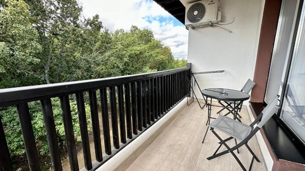 Uma varanda com guarda-corrimão preto, uma mesa com cadeiras e uma janela com vista para árvores.