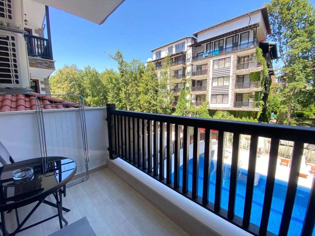A balcony with a glass table and chairs, overlooking a swimming pool and adjacent buildings.