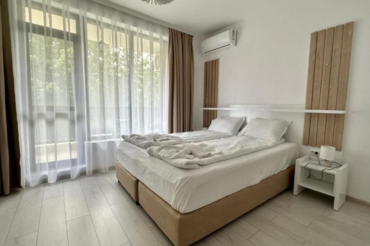 A bedroom with a bed, a bedside table, an air conditioning unit, and a window with curtains.