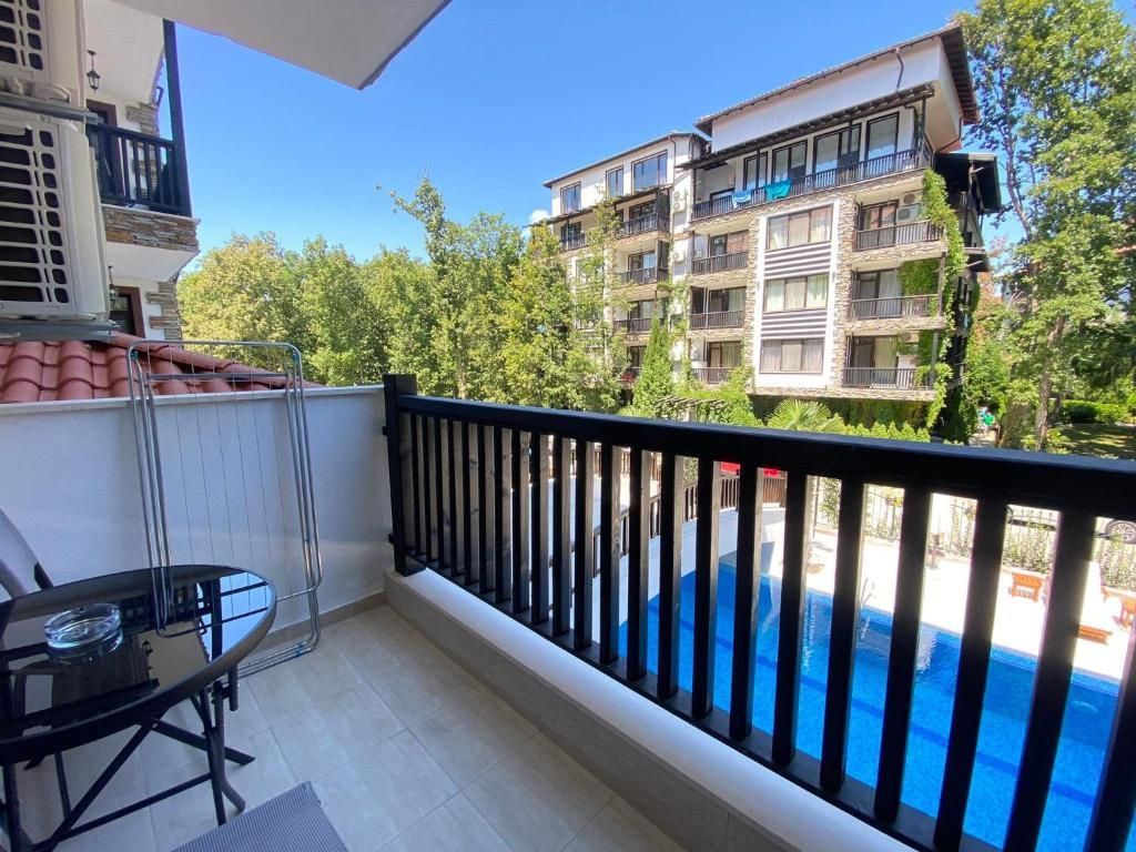 A balcony with a glass table and chairs, a pool in the background, and a view of a multi-story building with balconies.