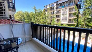 A balcony with a glass table and chairs, a pool in the background, and a view of a multi-story building with balconies.