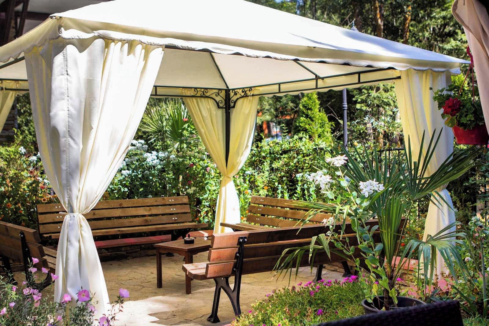 Uma área de estar coberta ao ar livre, com bancos e uma mesa, rodeada por vegetação exuberante e flores.