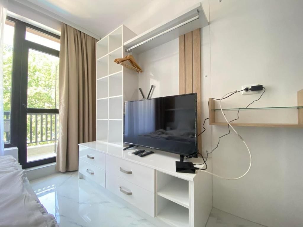 A modern bedroom with a TV on a white cabinet, a bed, and a balcony with curtains.