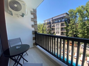 A balcony with a table and chairs, a pool in the background, and a building with multiple balconies.