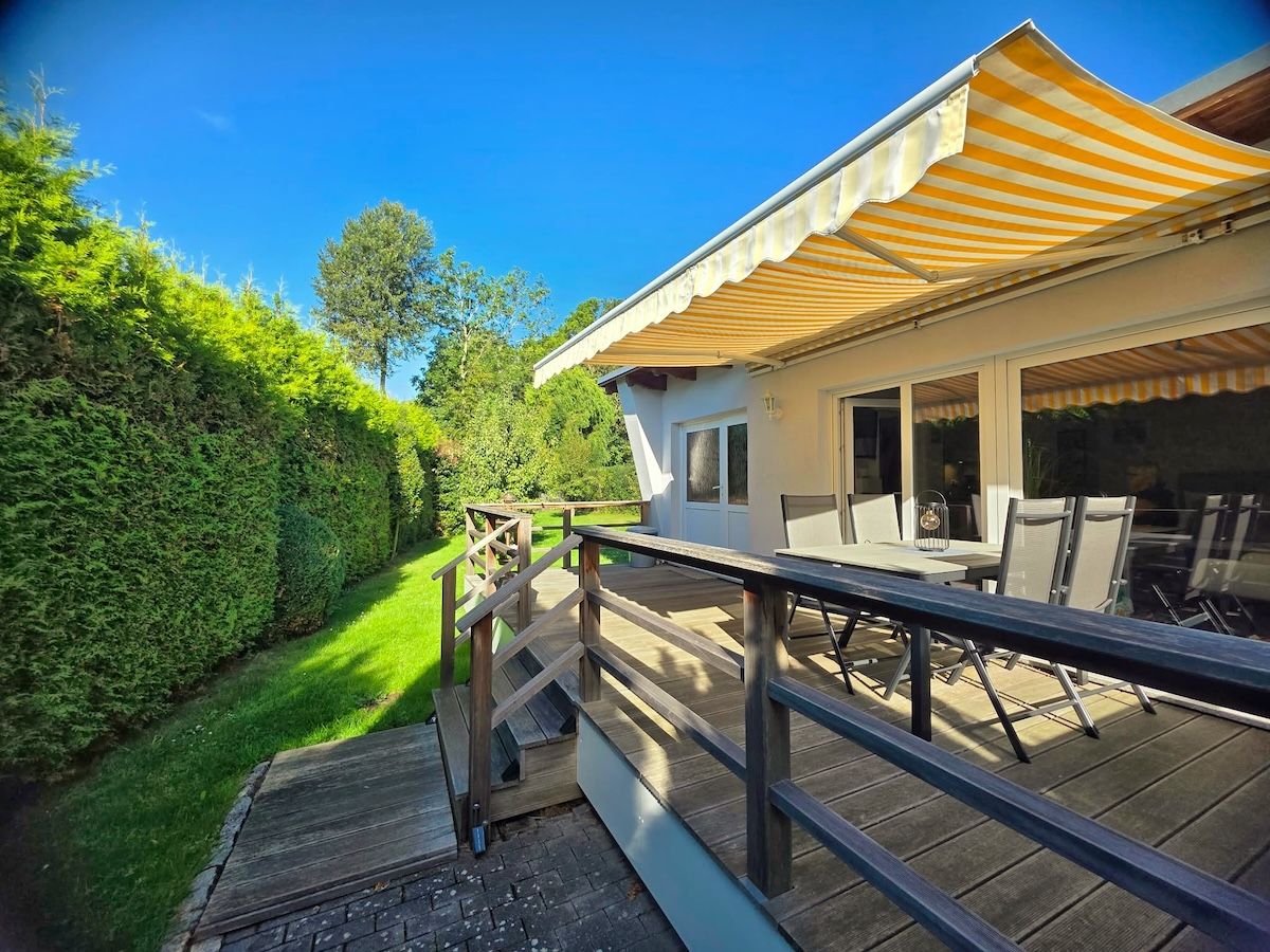 Une terrasse en bois avec une rambarde, un espace extérieur aménagé et abrité, et une vue sur un jardin bien entretenu.