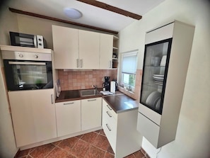 Eine kleine Küche mit weißen Schränken, einem eingebauten Backofen, einer Kaffeemaschine und einem Fenster mit Jalousien.