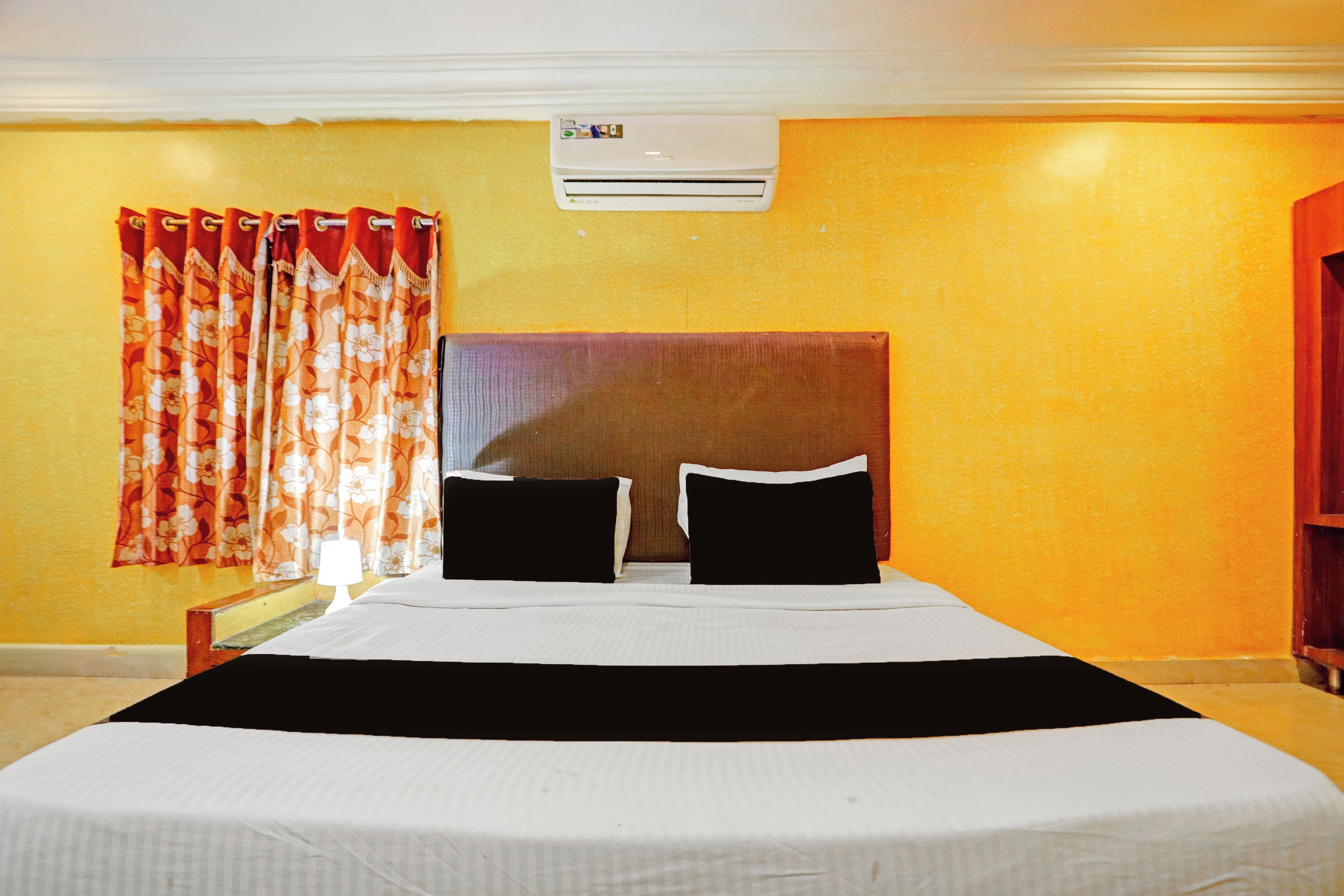 Habitación de hotel con una cama, dos almohadas, una mesita de noche, una lámpara, una ventana con cortinas y un equipo de aire acondicionado en la pared.