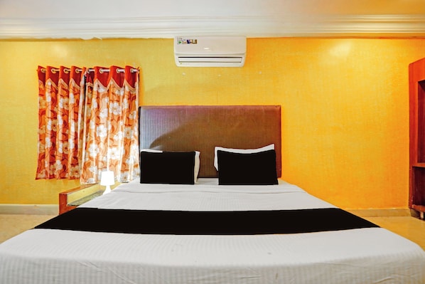 A hotel room with a bed, two pillows, a nightstand, a lamp, a window with curtains, and an air conditioning unit on the wall.