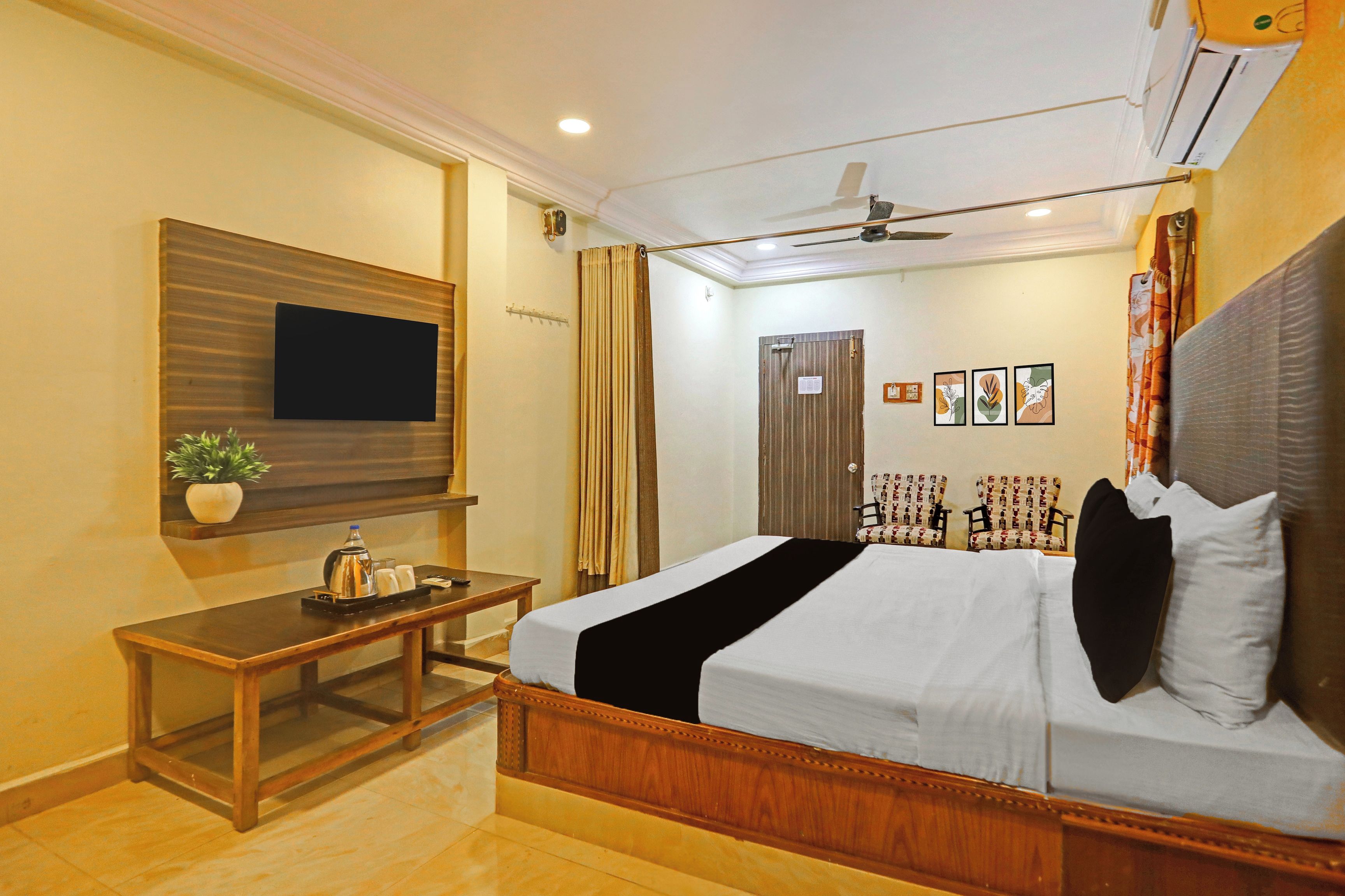 A hotel room with a bed, a TV mounted on the wall, a desk with a plant, and a ceiling fan.