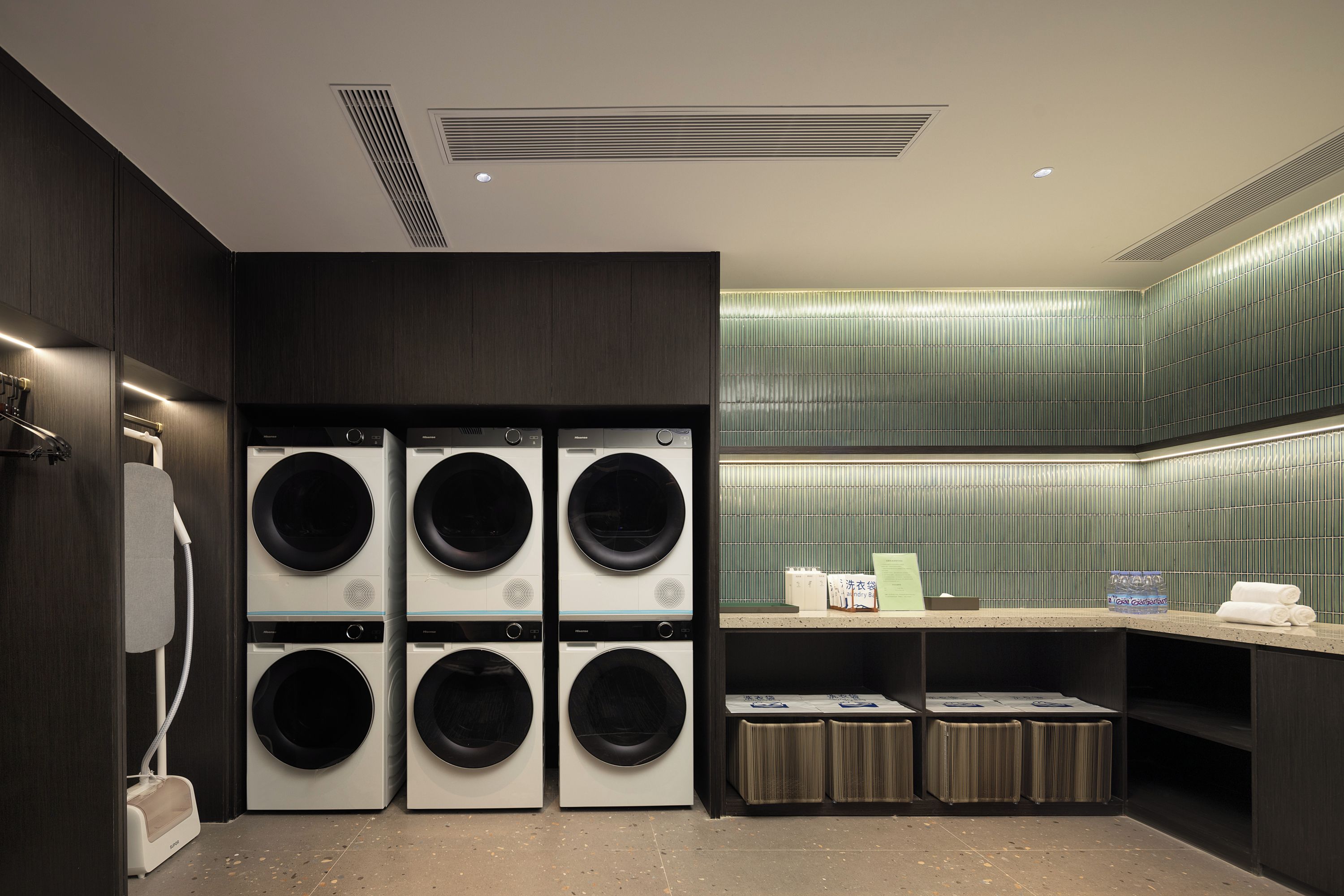 A modern laundry room with six front-loading washing machines, a drying rack, and a countertop with a water dispenser.