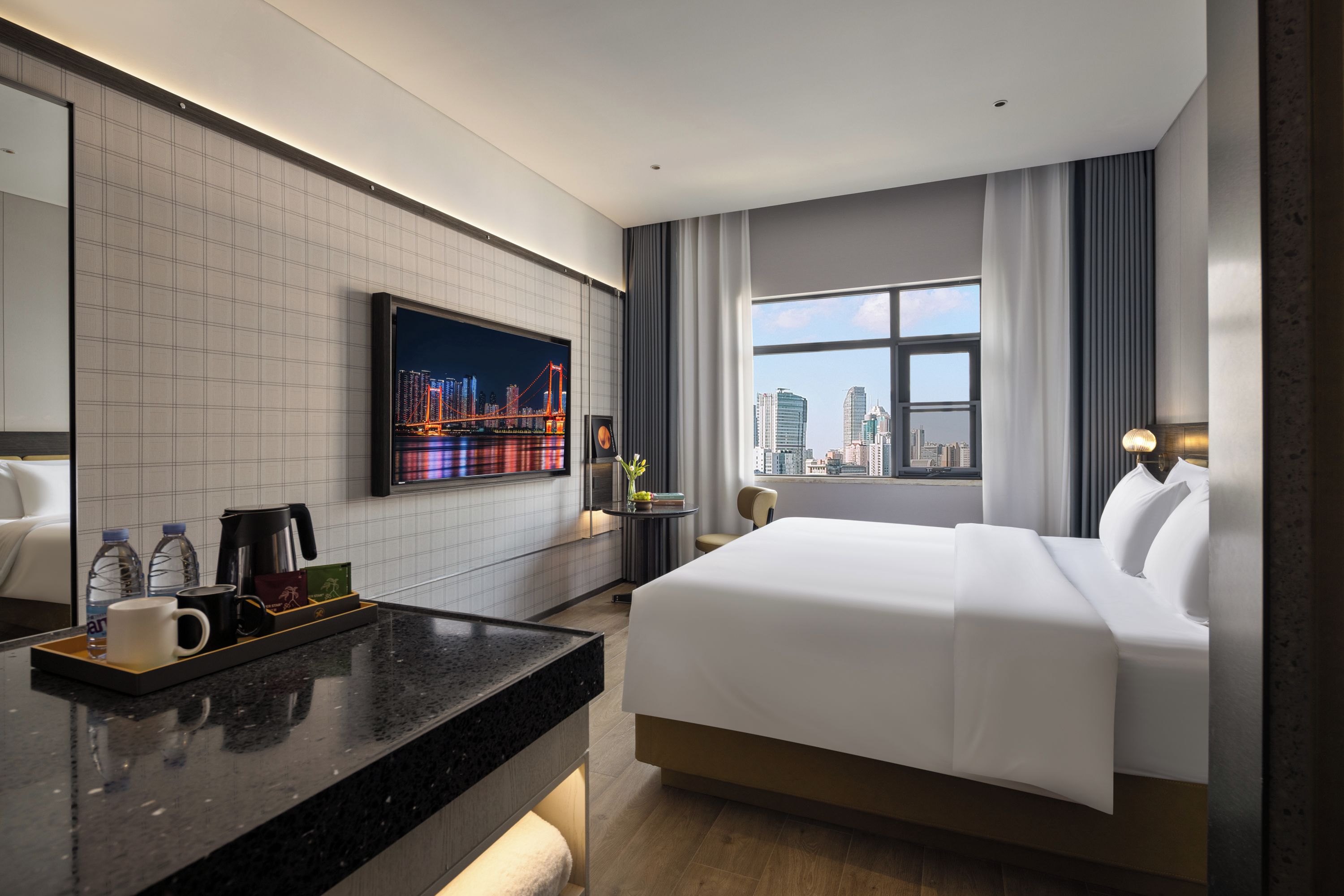 A hotel room with a large bed, a flat-screen TV mounted on the wall, a city view through the window, a small table with a tray of items, and a modern bathroom.