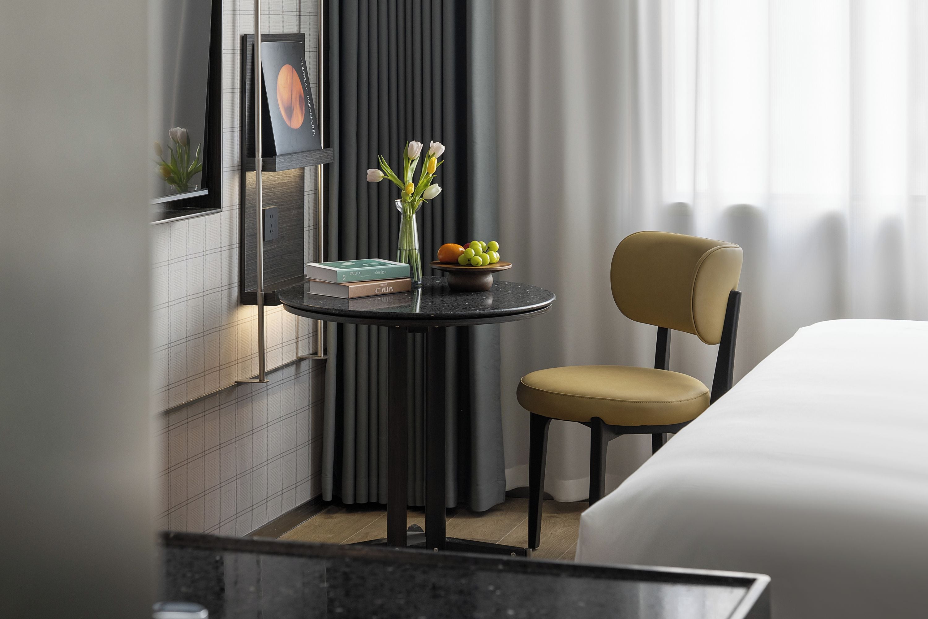 A modern hotel room with a round table, a chair, a bed, a small table with books and a vase of flowers, and a window with curtains.